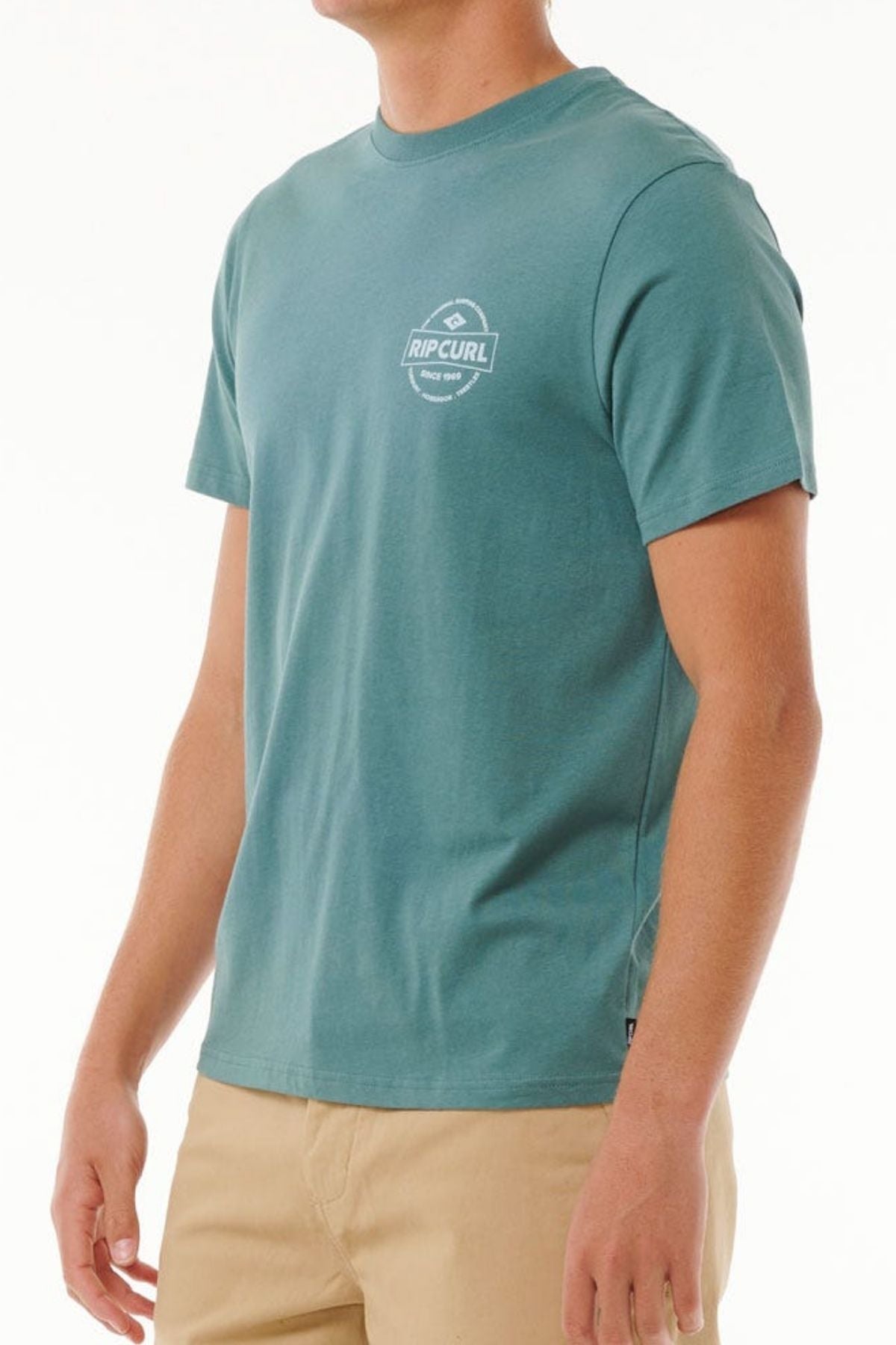 A man wears the Rip Curl Staple Tee in teal, made from soft cotton jersey with a small white logo on the left chest, paired with tan pants and standing against a plain white background.