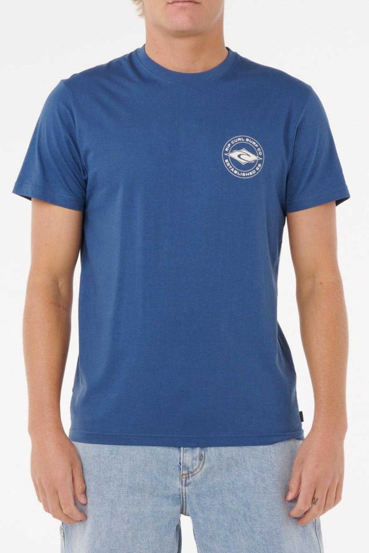 A person wearing a blue Rip Curl Staple Tee with a small white circular logo on the left chest and light blue jeans stands against a plain white background. The Rip Curl Staple Tee is made from cotton for comfortable everyday style.