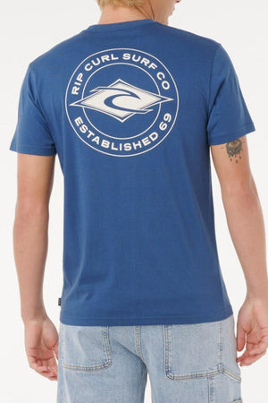 A person wearing the Rip Curl Staple Tee in blue, featuring a large white "Rip Curl Surf Co Established 69" logo on the back, is paired with light blue denim shorts. The comfortable cotton t-shirt is shown from behind.