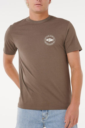 Wearing the Rip Curl Staple Tee, a brown cotton t-shirt with a small circular logo on the left chest, and light blue jeans, someone stands in front of a plain white background.