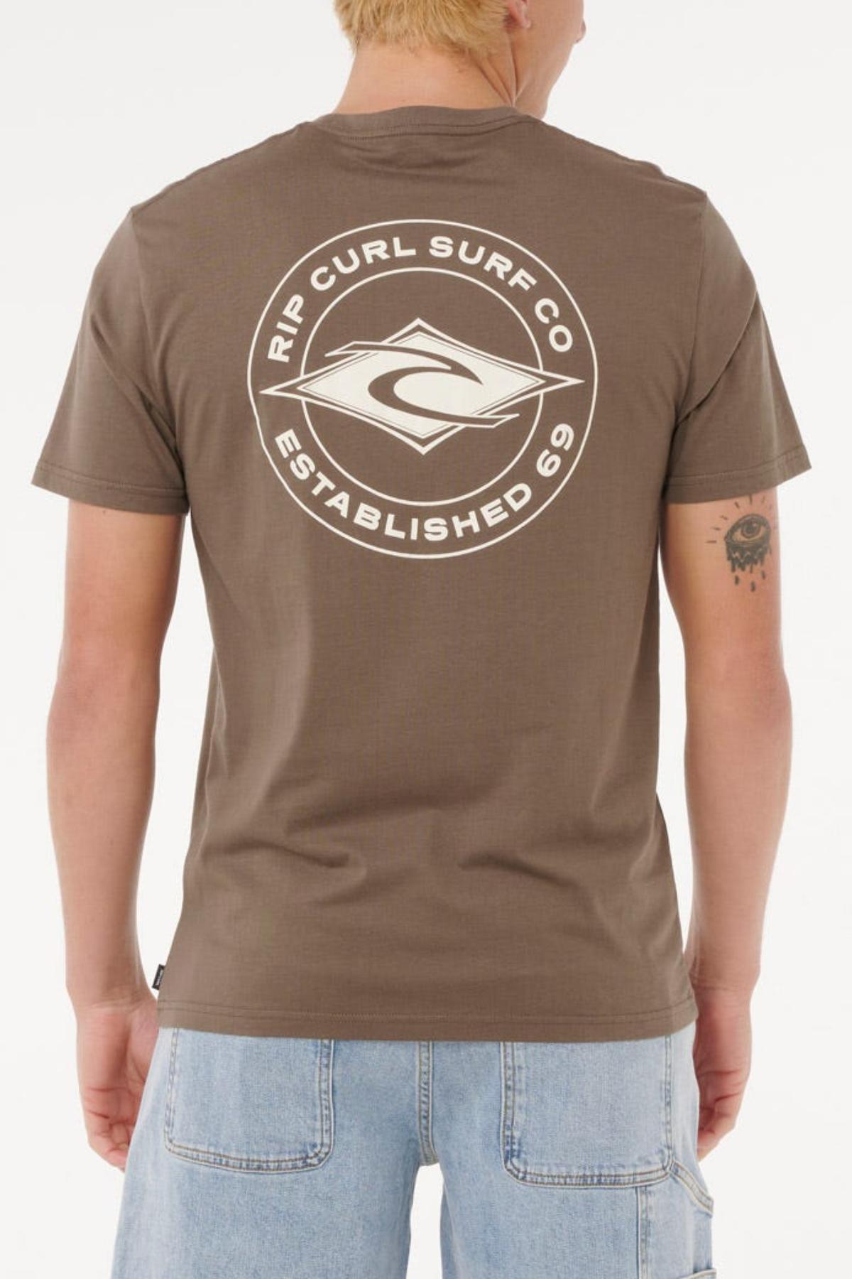 A person with short blond hair wears a Rip Curl Staple Tee—cotton, comfortable fit, with a large white "Rip Curl Surf Co Established 69" logo on the back—paired with light blue denim shorts.