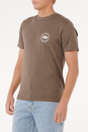 A person wears the brown Rip Curl Staple Tee, featuring a small circular white logo on the left chest, paired with light blue jeans and standing against a plain white background. The cotton tee offers a comfortable fit.