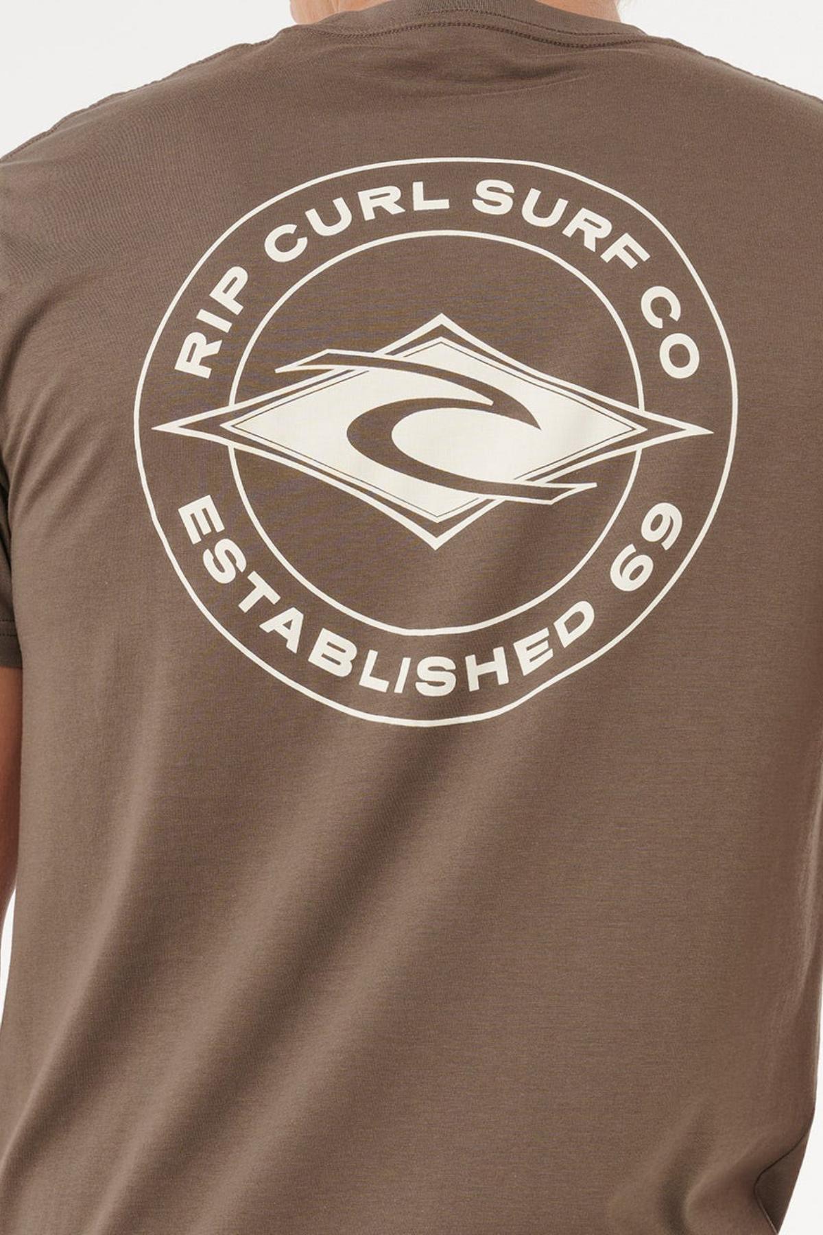 Someone wearing the Rip Curl Staple Tee in brown, featuring a large white Rip Curl Surf Co. logo, "Established 69" text, and stylized diamond-shaped waves on the back. This cotton t-shirt ensures a comfortable fit.
