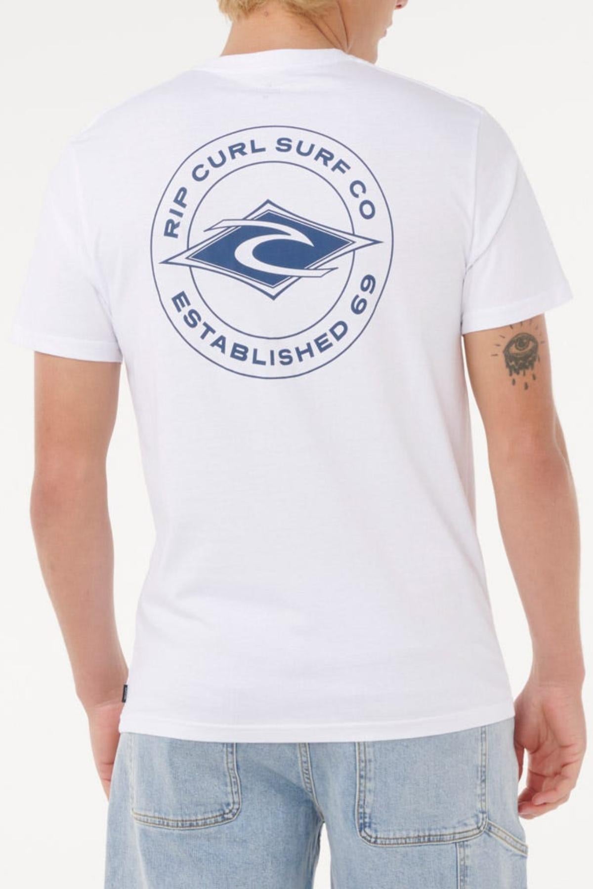 A person wears a Rip Curl Staple Tee, a white cotton jersey with a large blue back logo, styled with light blue jeans, short blonde hair, and a tattoo on their left arm.