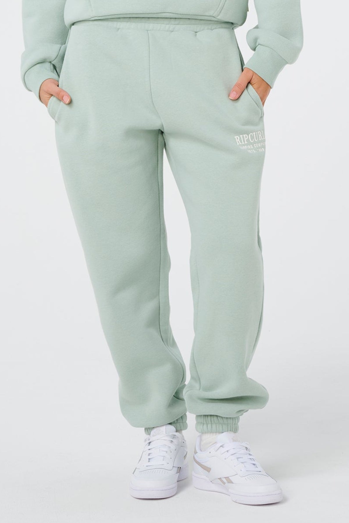 A person wears light green Rip Curl Surf Staple Trackpant with hands in pockets and white sneakers. "RIP CURL" is printed on the left thigh. The background is plain white.