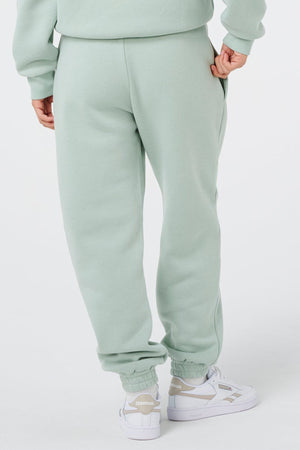 A person stands with their back to the camera in a light green Rip Curl Surf Staple Trackpant and matching sweatshirt, paired with white Reebok sneakers. The plain white background highlights the cozy loungewear look.
