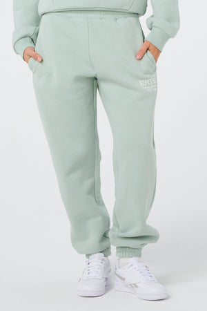 A person wears light green Rip Curl Surf Staple Trackpant with hands in pockets and white sneakers. "RIP CURL" is printed on the left thigh. The background is plain white.