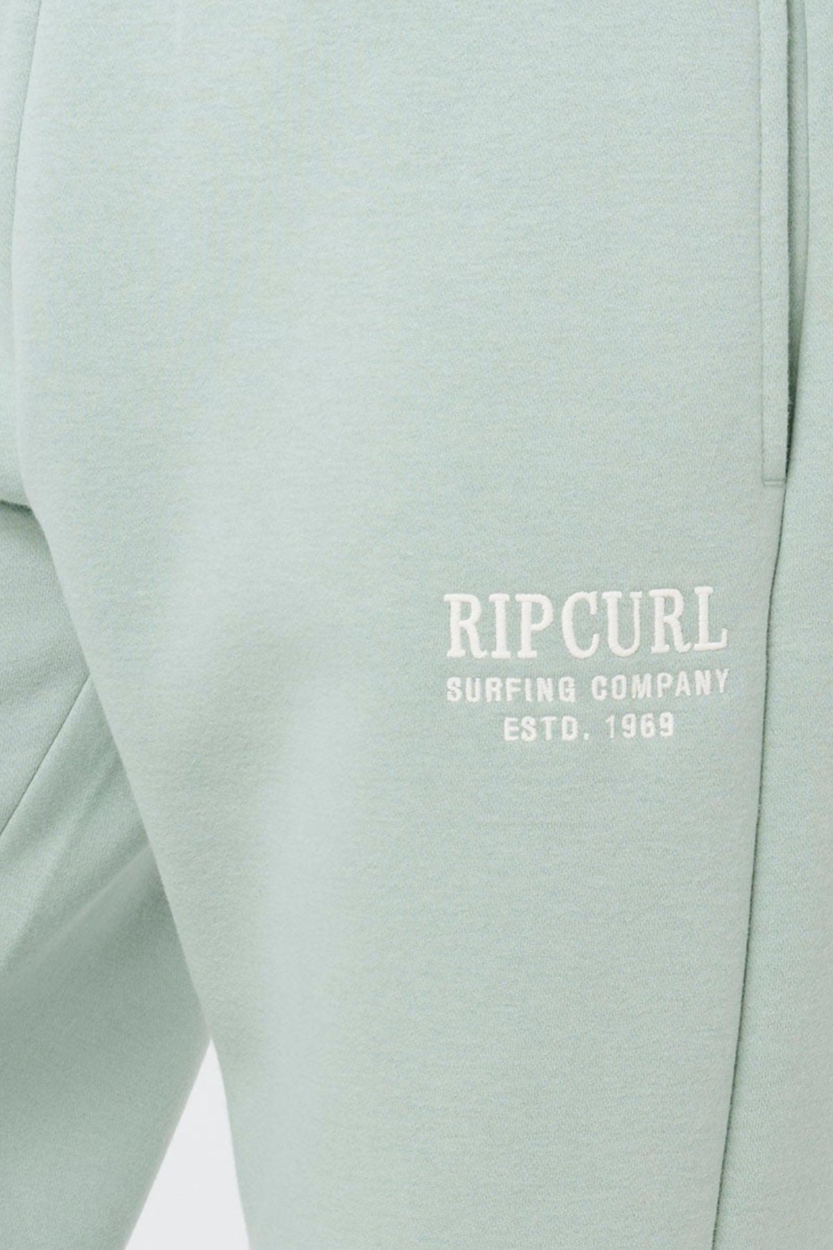 Close-up of light green Rip Curl Surf Staple Trackpant with a pocket and white "RIP CURL SURFING COMPANY ESTD. 1969" text on the upper thigh—ideal, comfy loungewear for a relaxed day.