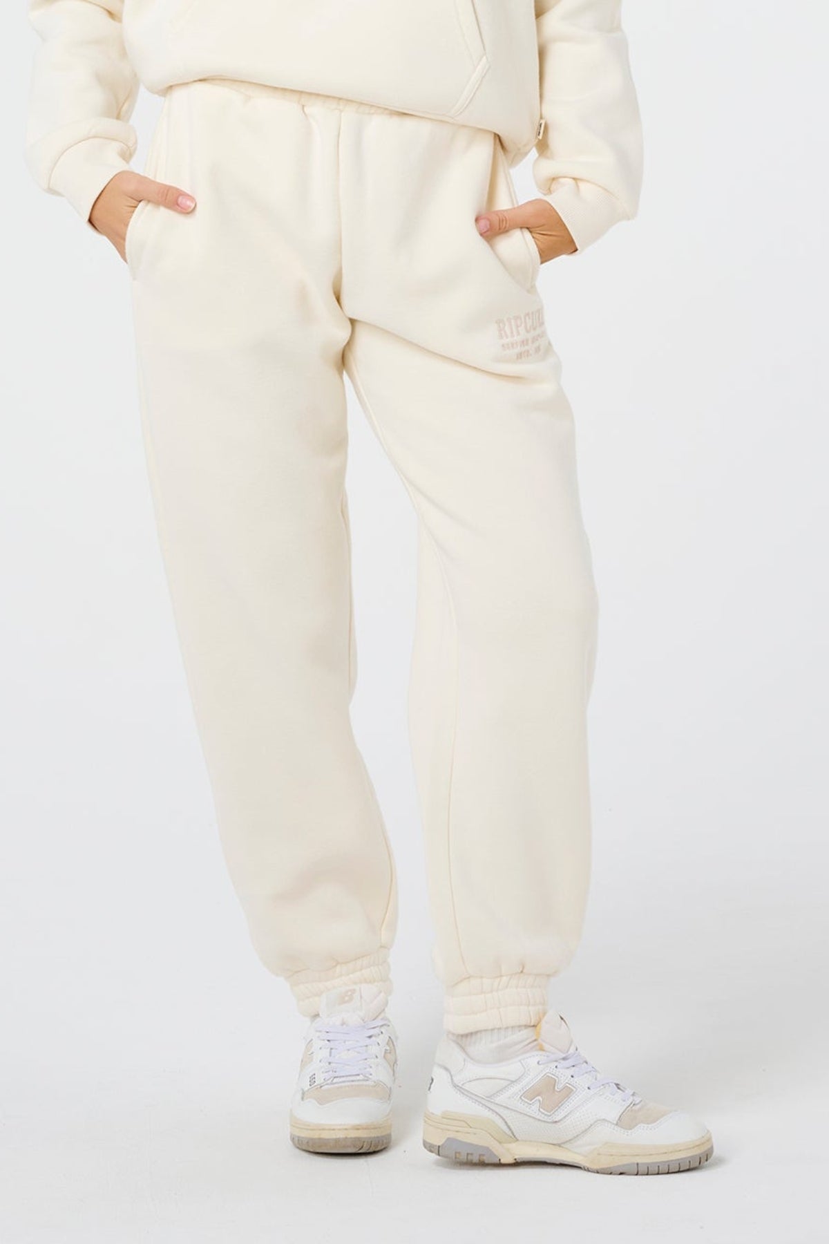 Wearing Rip Curl Surf Staple Trackpant and matching sneakers, a person stands hands in pockets against a white background, embodying a relaxed surf vibe.