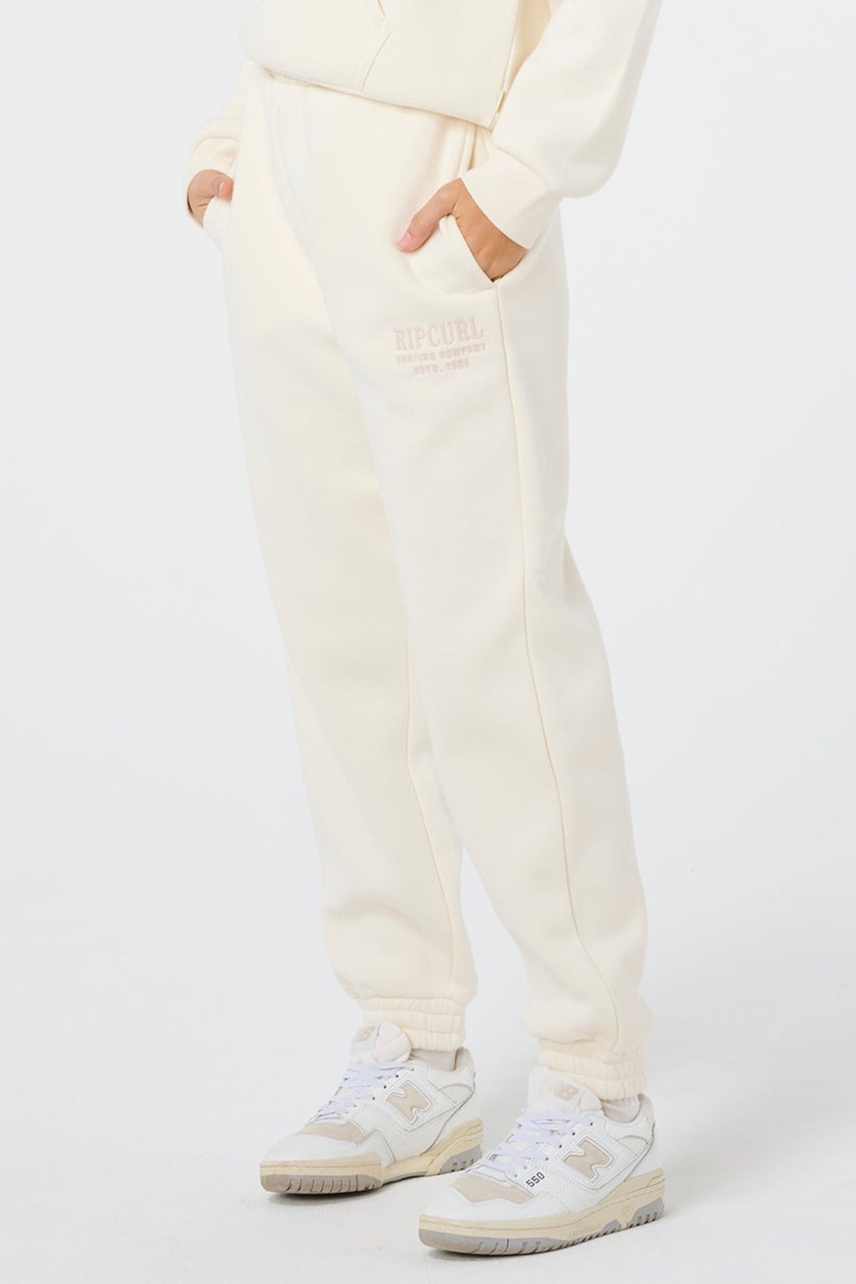 Wearing off-white Rip Curl Surf Staple Trackpants and white sneakers, a person stands with hands in pockets against a plain white background. Only the lower half is visible, highlighting a relaxed surf-inspired style.