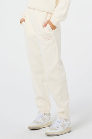 Wearing off-white Rip Curl Surf Staple Trackpants and white sneakers, a person stands with hands in pockets against a plain white background. Only the lower half is visible, highlighting a relaxed surf-inspired style.