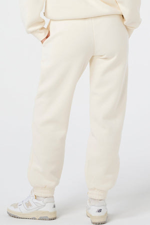 A person stands with one hand in their pocket, shown from the back, wearing Rip Curl Surf Staple Trackpants and a matching cream sweatshirt with white New Balance sneakers for a relaxed surf-inspired look.
