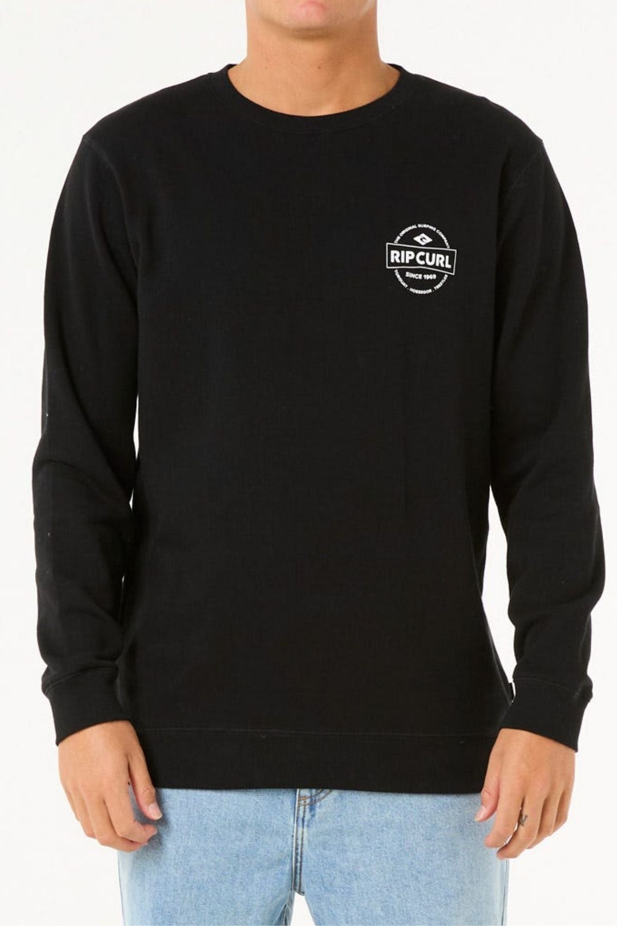 A person stands against a white background, wearing the black Rip Curl Stapler Crew sweatshirt—crafted from cotton polyester fleece with a small white logo on the upper left chest—and light blue jeans.