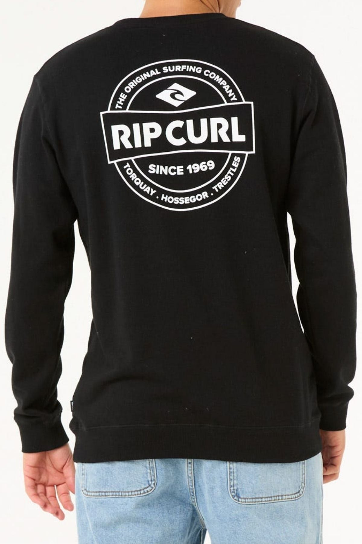 A person wears light blue jeans and a black Rip Curl Stapler Crew sweatshirt, crafted from cotton polyester fleece, featuring a white circular logo and text on the back for unique style and comfort.