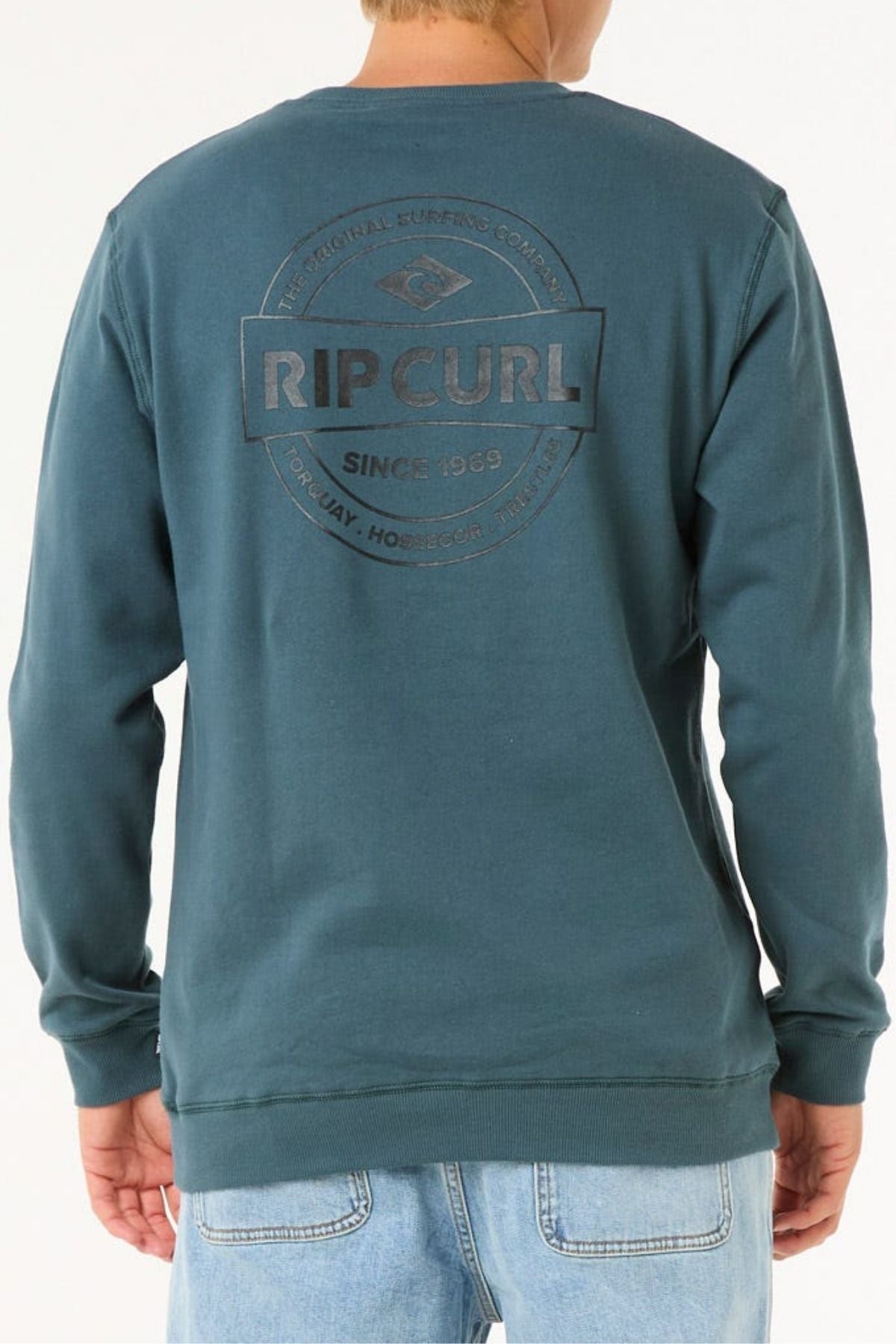 A person seen from behind wears a teal Rip Curl Stapler Crew sweatshirt with a circular logo and "RIP CURL SINCE 1969" on the back, paired with light blue jeans—ideal for adventures.