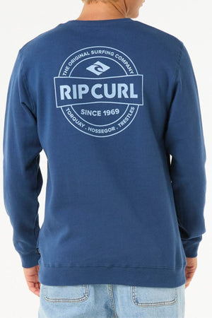 A person wearing the Rip Curl Stapler Crew in blue is seen from the back. This cotton-polyester fleece sweatshirt features unique front and back prints, including a large Rip Curl logo and "The Original Surfing Company" text.