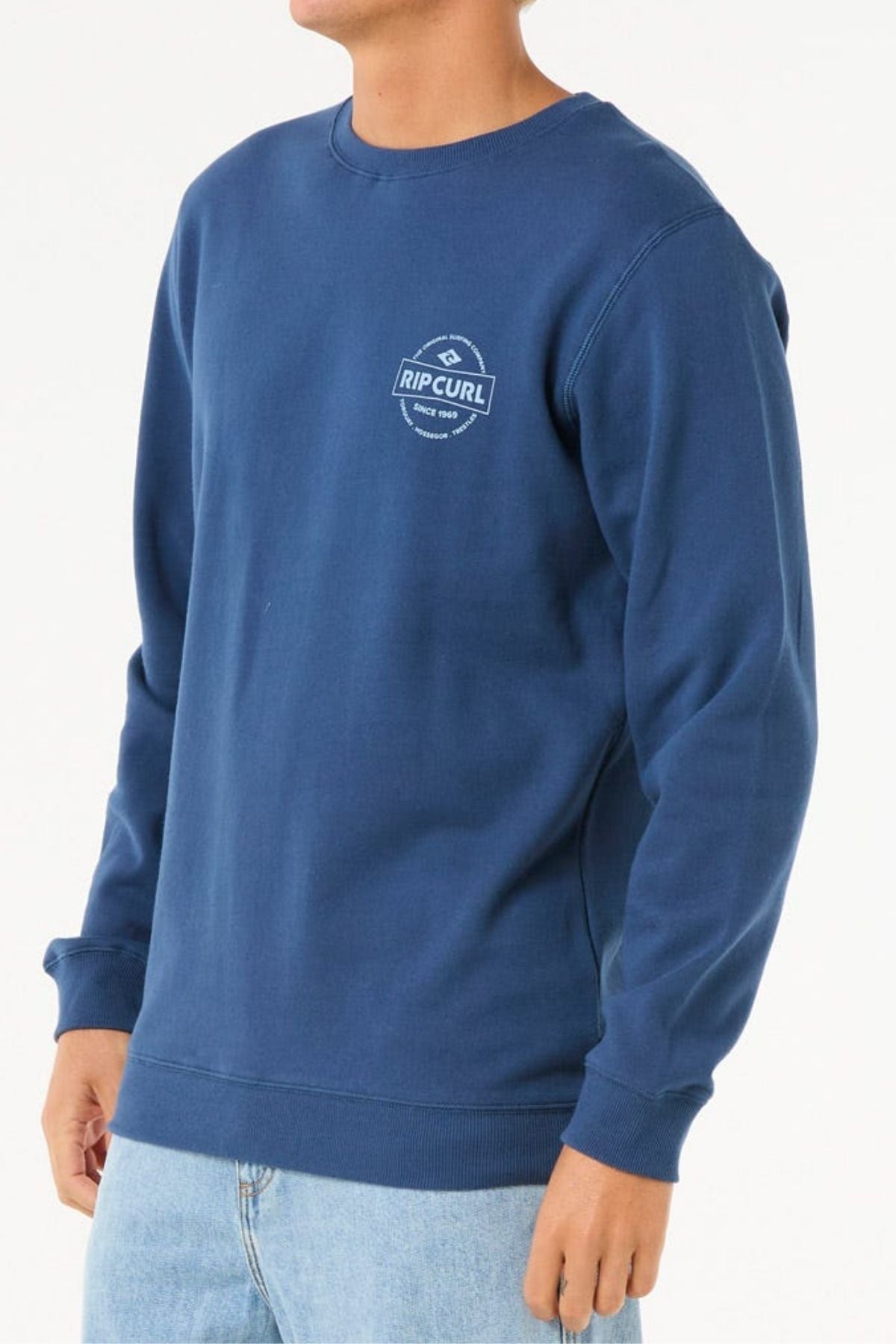 A person wearing a blue Rip Curl Stapler Crew sweatshirt and light blue jeans stands against a plain white background. The sweatshirt has a small Rip Curl logo on the left chest.