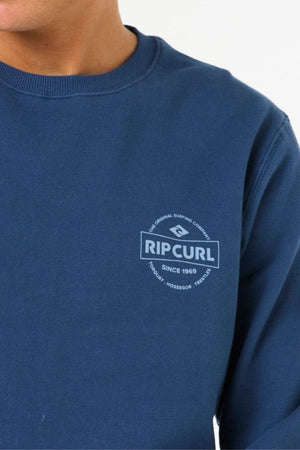 A person wearing a blue Rip Curl Stapler Crew fleece sweatshirt featuring unique front and back prints with the white "Rip Curl, Since 1969, The Original Surfing Company" logo and text.