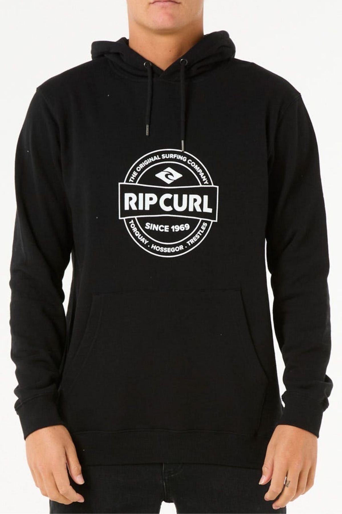Someone wearing the Rip Curl Stapler Hood, a black cotton polyester fleece hoodie featuring a white "Rip Curl" logo plus "The Original Surfing Company," "Since 1969," and "Proudly - Hossegor - Torquay." Their face is partially obscured.
