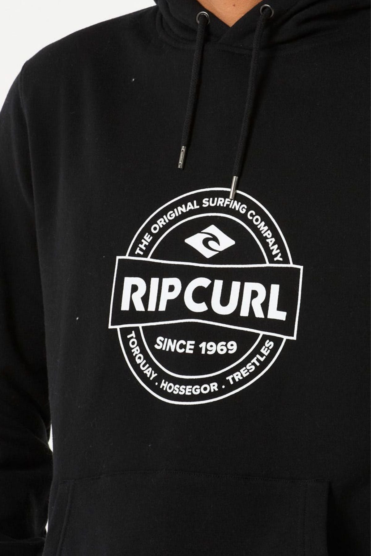 Close-up of a person wearing the Rip Curl Stapler Hood, a black fleece hoodie with a white "Rip Curl" logo and "The Original Surfing Company Since 1969 Torquay Hossegor Trestles" text.