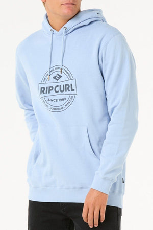 Wearing the Rip Curl Stapler Hood in light blue, made from a cotton-polyester fleece blend with a large chest logo, long sleeves, drawstring hood, and front pocket. The wearer’s face is partially out of frame.