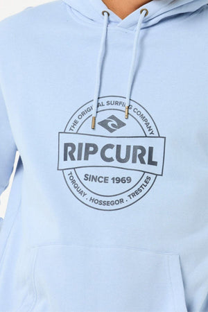 Someone wearing the Rip Curl Stapler Hood—a light blue cotton-polyester fleece hoodie—features a circular logo with "RIP CURL The Original Surfing Company Since 1969" and place names Torquay, Hossegor, and Trestles on the front.