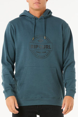 A person wears the Rip Curl Stapler Hood in teal, made from soft cotton-polyester fleece. It features a front pocket, drawstrings, and the black Rip Curl logo with "Since 1969" on the chest. Shown from shoulders to hips.