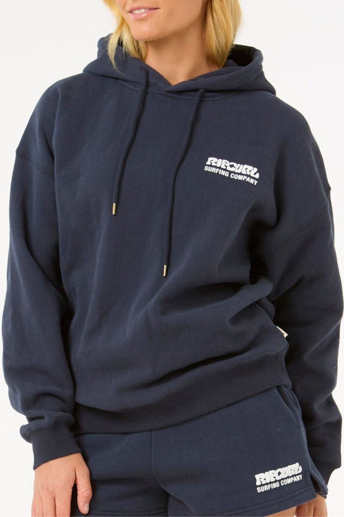 A person wears a navy blue Rip Curl Surf Puff Heritage Hood and matching shorts, both featuring "RIP CURL SURFING COMPANY" in white, set against a plain background.