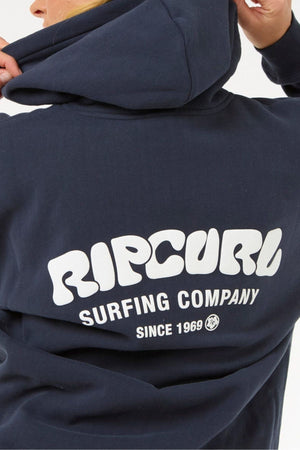 A person wears a dark blue Rip Curl Surf Puff Heritage Hood sweatshirt with the hood up. The back displays white text: "RIP CURL SURFING COMPANY SINCE 1969.