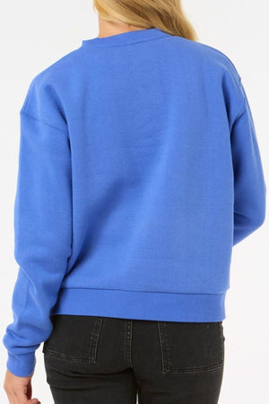 A person with long blonde hair, shown from the back against a plain light background, is wearing the Rip Curl Surf Staple Relaxed Crew in bright blue with black jeans.