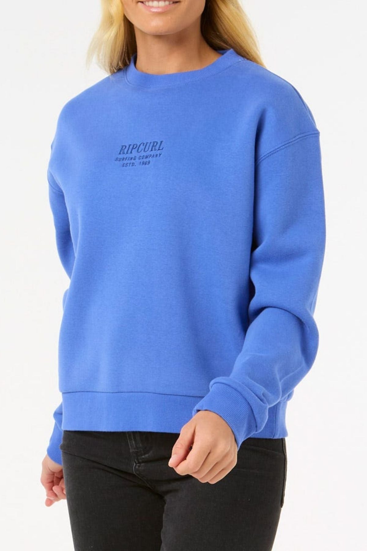 A person in a bright blue Rip Curl Surf Staple Relaxed Crew sweatshirt and black pants smiles while standing against a plain white background.