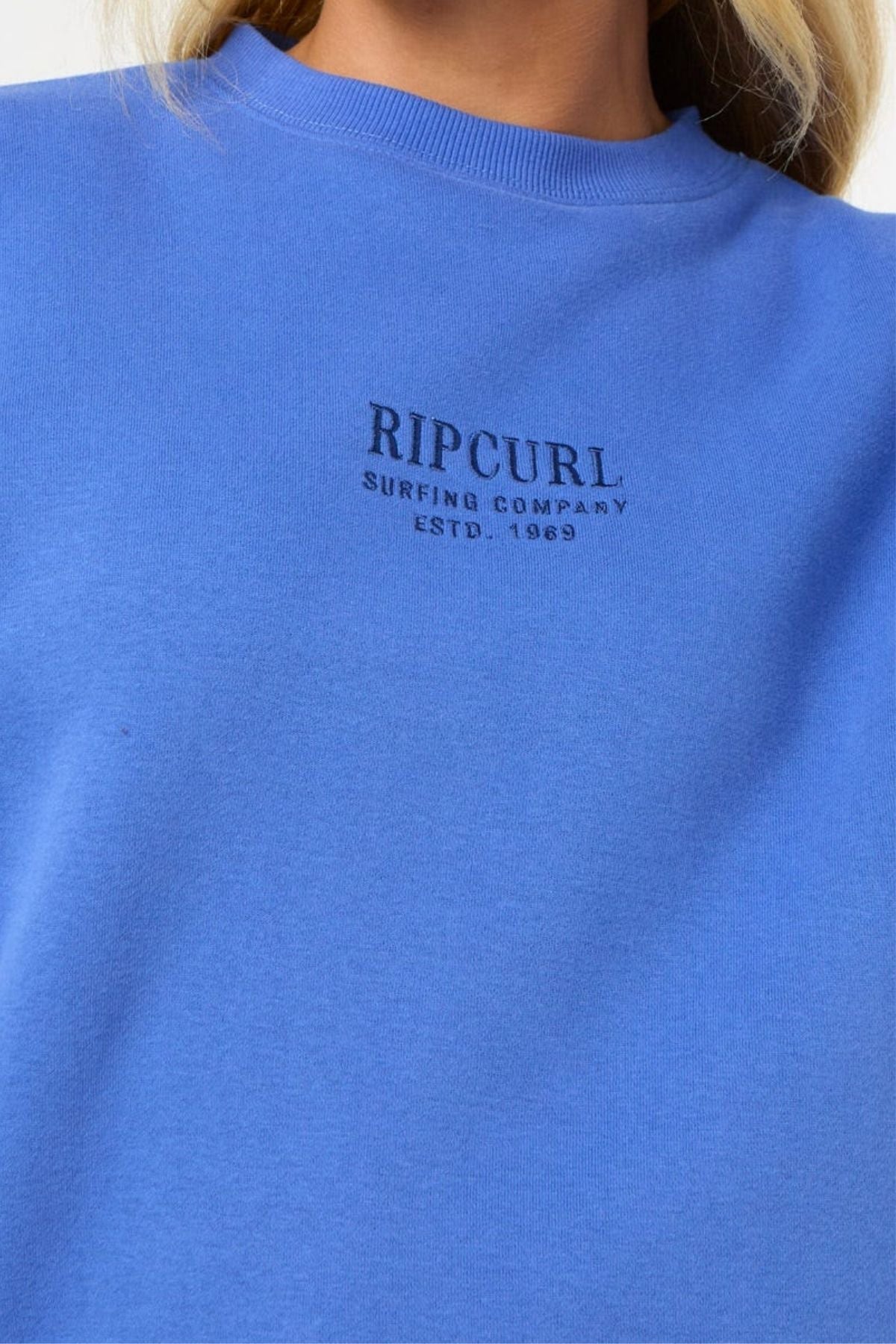 Close-up of a person wearing the blue Rip Curl Surf Staple Relaxed Crew, made from soft cotton polyester fleece, with "RIP CURL SURFING COMPANY ESTD. 1969" in black on the front. Only the lower face and upper chest are shown.