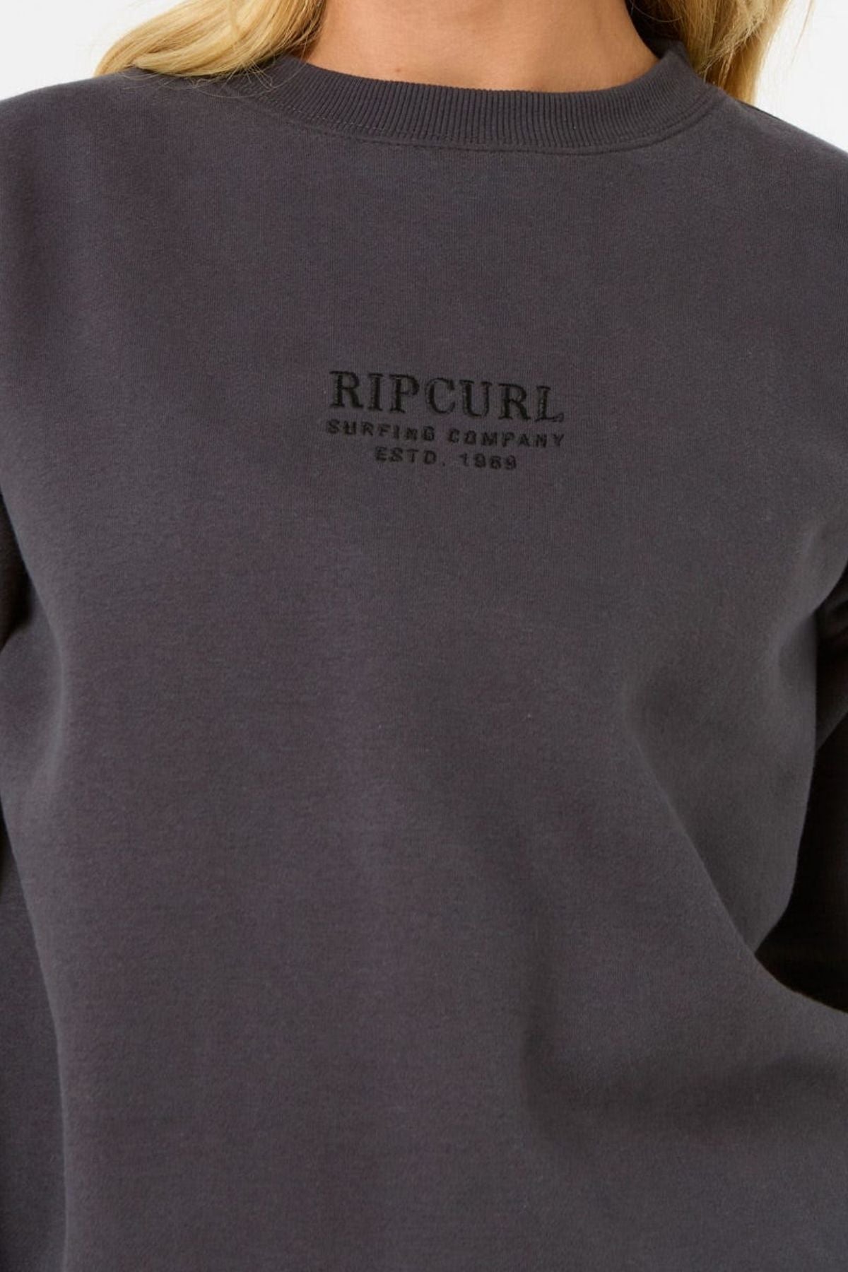 A close-up shows a person wearing the Rip Curl Surf Staple Relaxed Crew in dark gray fleece, featuring "RIP CURL Surfing Company Estd. 1969" embroidered on the chest. Only their lower face and upper torso are visible.