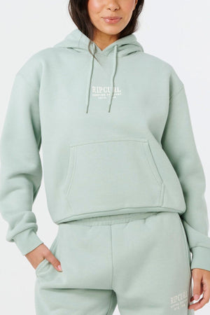 A woman wearing a light sage green Rip Curl Surf Staple Relaxed Hood and matching sweatpants stands with one hand in her pocket against a plain white background.