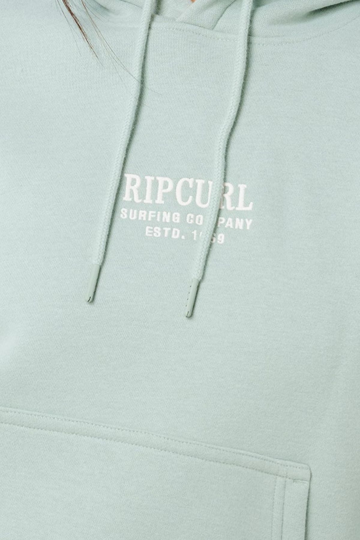 A close-up of the Rip Curl Surf Staple Relaxed Hood in light green, featuring white "RIP CURL SURFING COMPANY ESTD. 1969" text on the chest, a front pocket, drawstrings, and a relaxed fit—an essential surf pullover.