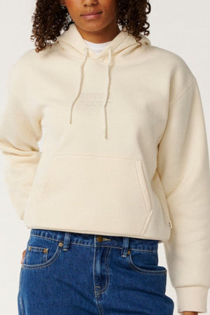 Wearing the Rip Curl Surf Staple Relaxed Hood in cream, styled with blue jeans, someone stands against a plain background. The hoodie features a relaxed pullover fit and subtle chest embroidery for a classic surf-inspired look.