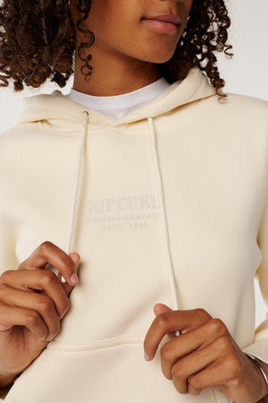 A person with curly hair wears a cream Rip Curl Surf Staple Relaxed Hood, "Surfing Company EST. 1969" embroidered on the chest, holding both hoodie strings. Only the lower half of their face is visible.