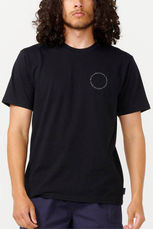 A person with long curly hair is wearing the Rip Curl Vaporcool Search Rubix Tee, a black T-shirt featuring a small, circular text design on the chest. Made with quick-dry, moisture-wicking fabric, it keeps you comfortable. They are standing against a plain background and wearing dark pants.