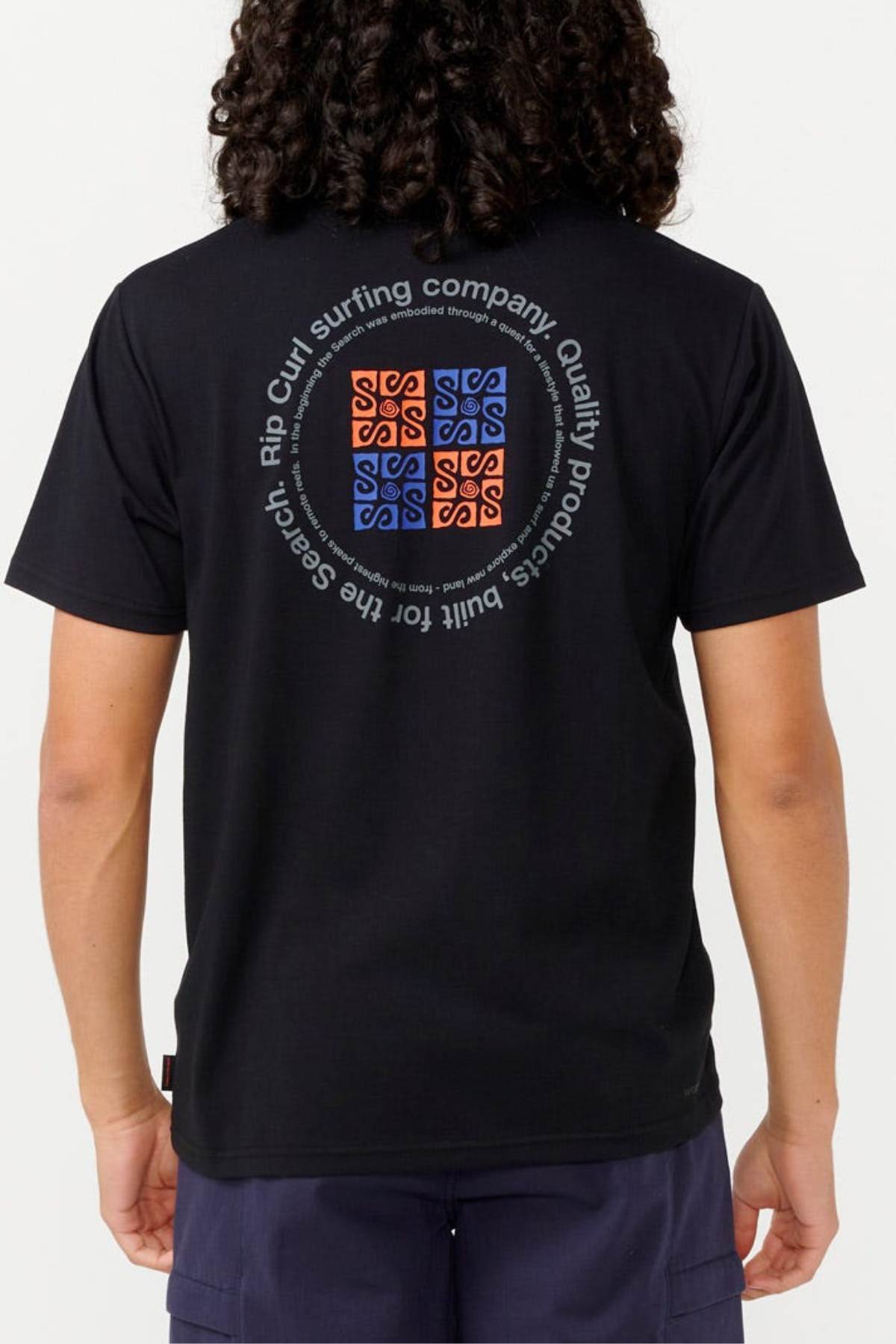 A person with long curly hair is wearing the Rip Curl Vaporcool Search Rubix Tee, which features an advanced cooling technology. The back of this black T-shirt displays a circular graphic design showcasing "Rip Curl surfing company" along with vibrant blue and orange square patterns. They are also sporting dark, quick-dry shorts.