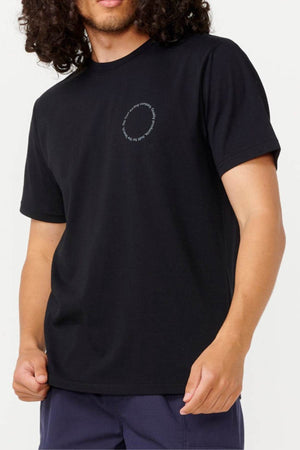 A person with curly hair is wearing the Rip Curl Vaporcool Search Rubix Tee in black, featuring a small circular text design on the chest and made from quick-dry material. They are also sporting dark blue pants with VaporCool technology against a plain white background.