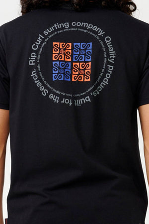 Back view of a person wearing the black Rip Curl Vaporcool Search Rubix Tee. Crafted with quick-dry fabric, the shirt displays an orange and blue square design featuring abstract patterns, surrounded by text that reads, "Rip Curl surfing company, Quality products, Built for the Search.