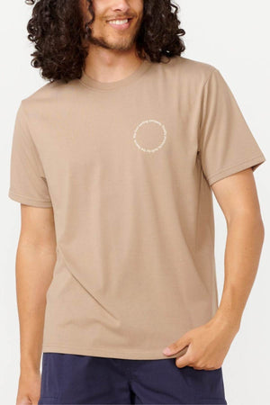 A person with long curly hair is wearing a Rip Curl Vaporcool Search Rubix Tee in beige, showcasing a circular text design on the chest. They pair it with dark blue pants, enjoying the comfort of the moisture-wicking fabric as they smile with one hand in their pocket.