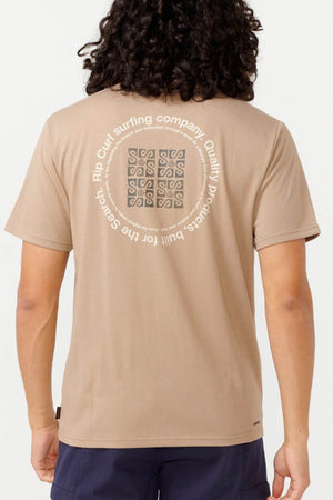 A person with long curly hair is wearing a beige Rip Curl Vaporcool Search Rubix Tee featuring a circular graphic on the back with the text, "Rip Curl surfing company" and "Quality Products." This VaporCool t-shirt provides moisture-wicking comfort. The individual is also dressed in dark shorts.