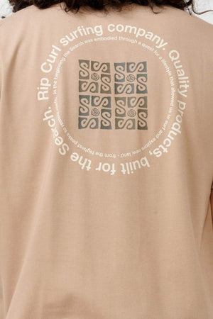 The Rip Curl Vaporcool Search Rubix Tee is a beige shirt that showcases a circular design on the back with abstract patterns at its center and text around it, stating "Rip Curl surfing company. Quality products built for the search." Made with VaporCool technology, it offers moisture-wicking and odor control to keep you fresh.