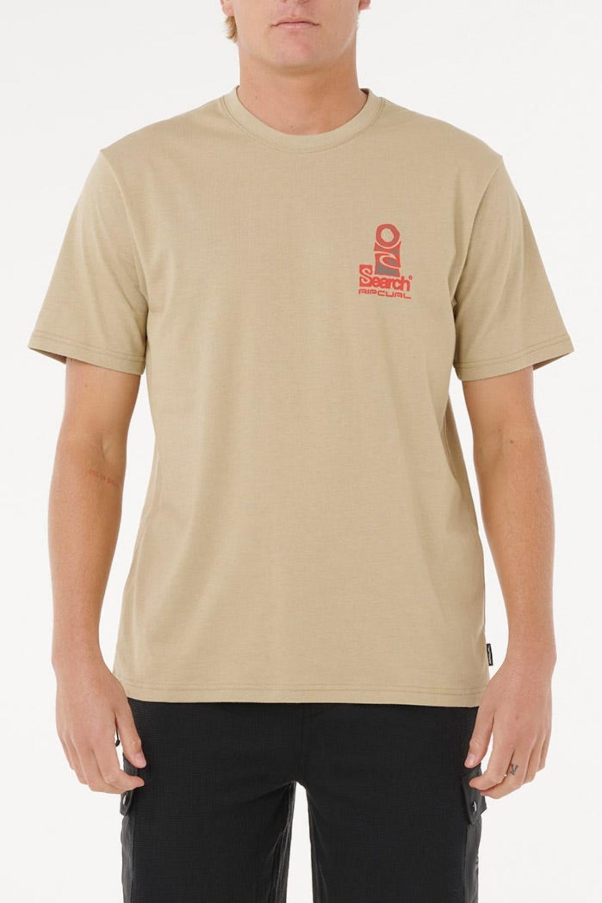 Someone stands against a plain white background wearing the beige Rip Curl Vaporcool Search Stack Tee, featuring moisture-control fabric, a small red "Search" graphic on the left chest, and black pants.