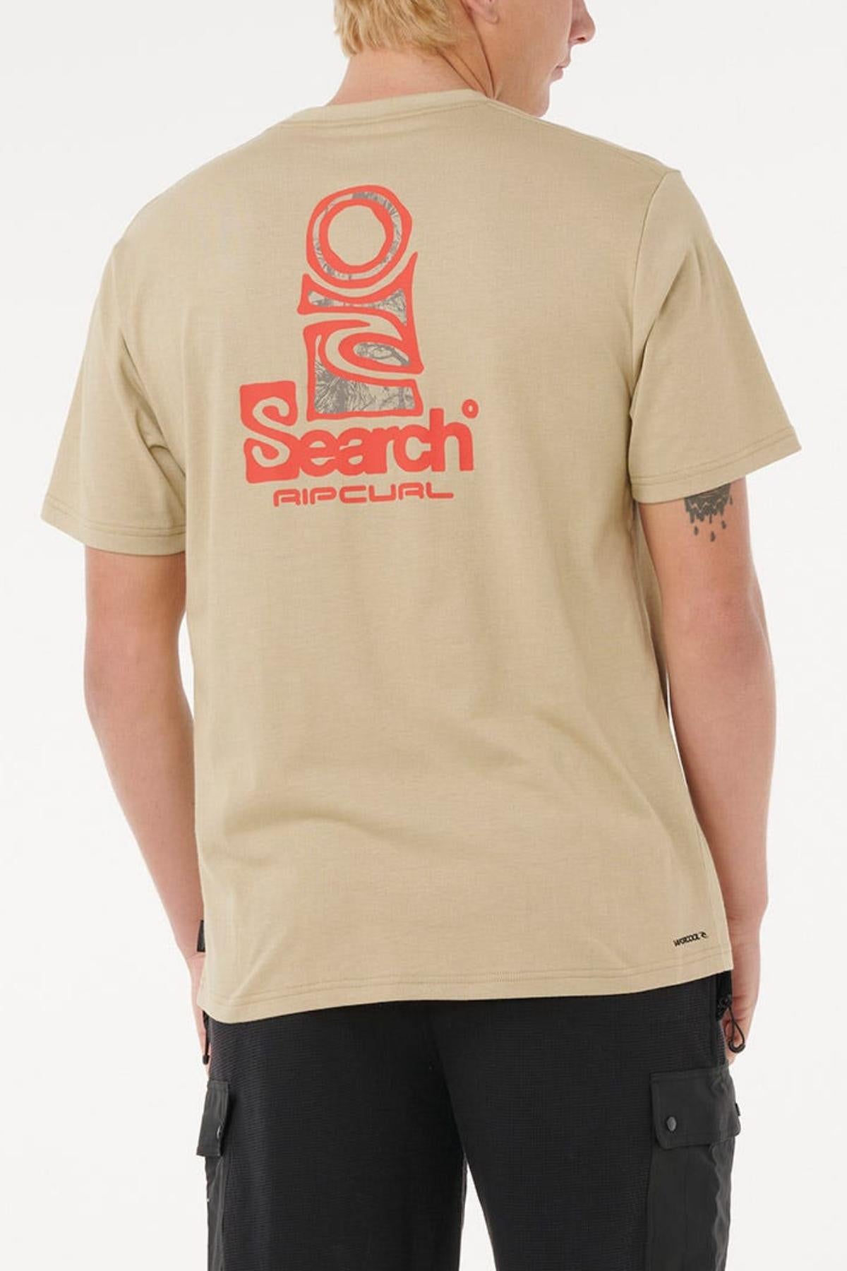 A person with short blond hair is seen from behind wearing a beige Rip Curl Vaporcool Search Stack Tee with a red "Search" and "Rip Curl" graphic on the back. They also have a small tattoo on their left arm.