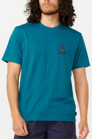 A person with long, curly hair stands against a plain background wearing the Rip Curl Vaporcool Search Stack Tee in teal, featuring a small logo on the chest and paired with dark pants, enjoying its quick-drying comfort.