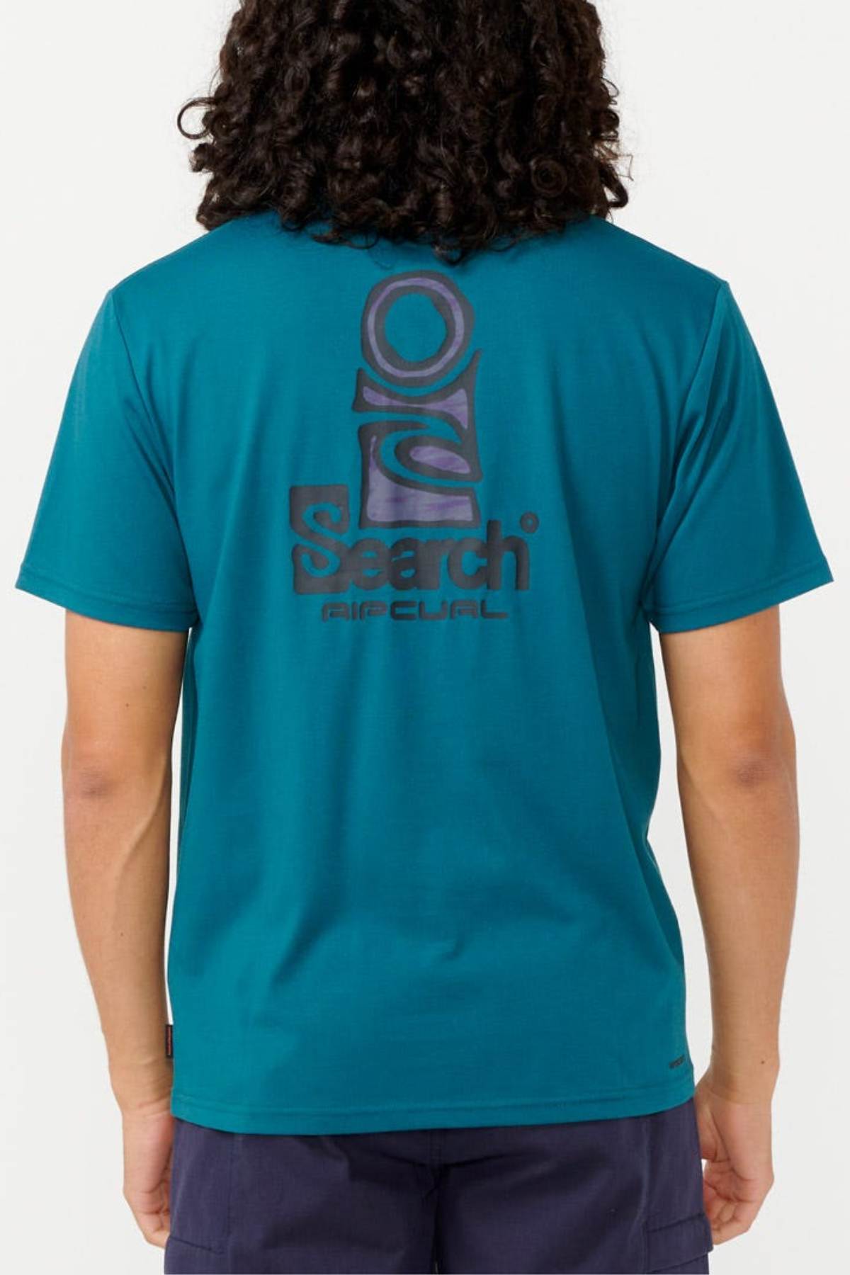 A person with long curly hair is wearing a teal Rip Curl Vaporcool Search Stack Tee, which features a large graphic and the words "Search Rip Curl" on the back. They are also dressed in dark shorts. The quick-drying clothing stands out against the plain white background.