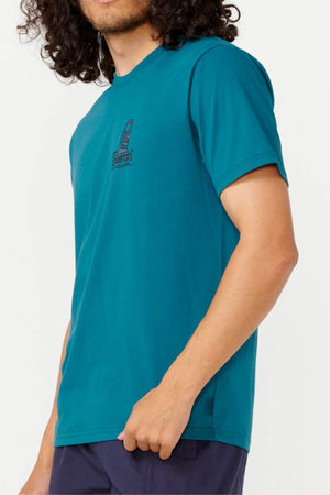 A person with curly hair is wearing the Rip Curl Vaporcool Search Stack Tee, a teal T-shirt featuring a small logo on the chest and designed with quick-drying technology. The image focuses on their torso, as it's cropped at the head and legs. This T-shirt is paired with navy pants, making it ideal for those busy days when comfort and ease are key.