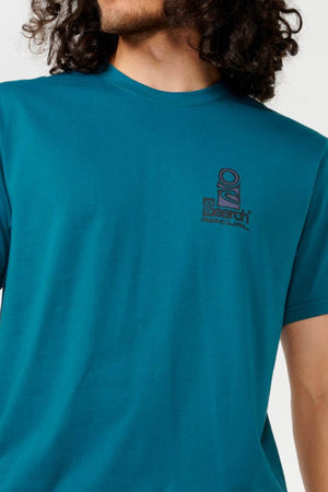 Someone wearing a Rip Curl Vaporcool Search Stack Tee in teal, featuring a small abstract design and text on the chest. Their long, curly hair flows as they turn slightly away from the camera, embodying style and innovation with quick-drying comfort.